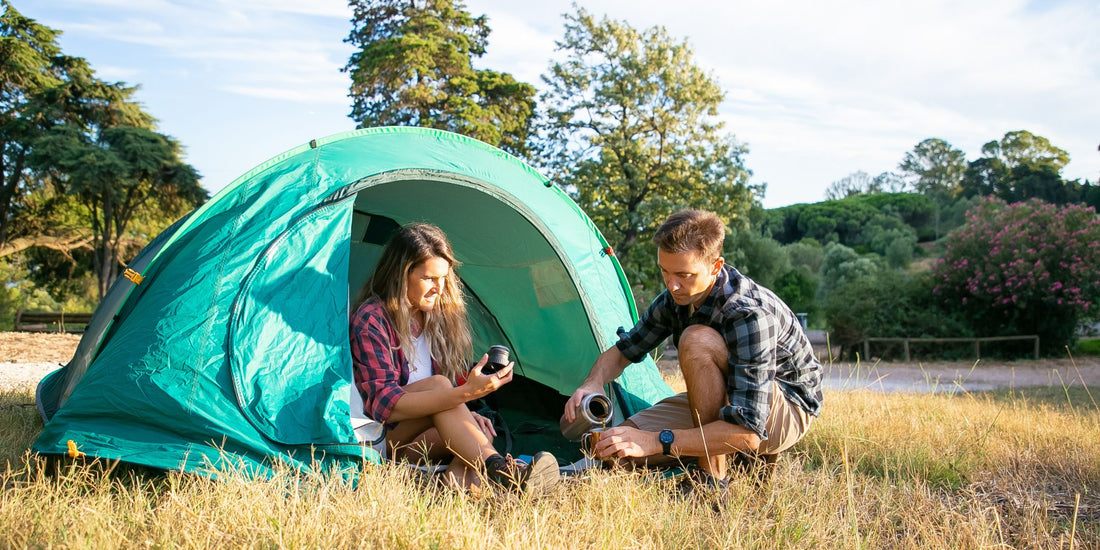 Young couple sitting outside green tent sharing drinks at campsite
