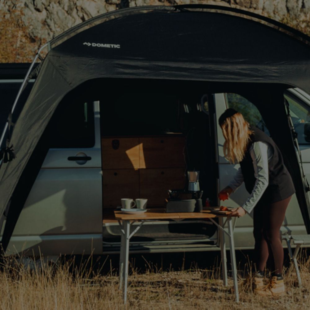 Woman preparing coffee at outdoor table beside Dometic van awning campsite